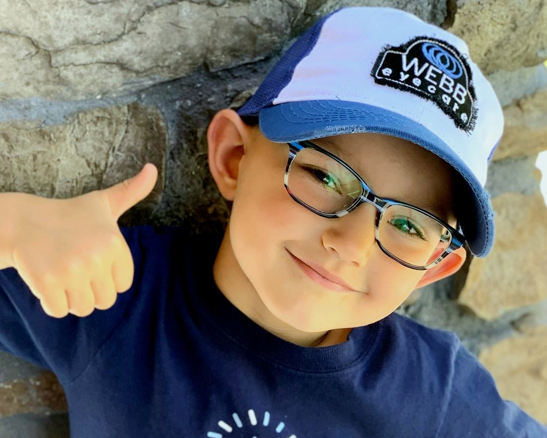 A young Webb Eyecare patient giving a thumbs up in his glasses and Webb cap.
